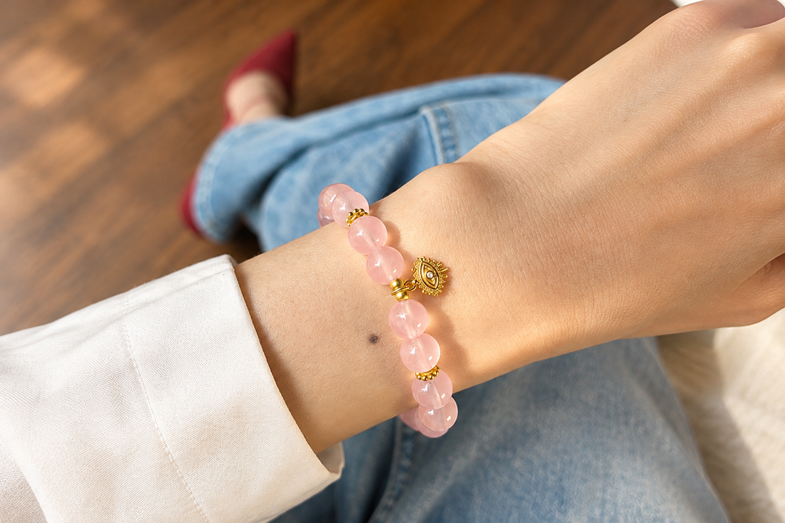 Pink rose quartz bead bracelet with a gold-tone evil eye charm worn on a wrist, styled with a white shirt and jeans.