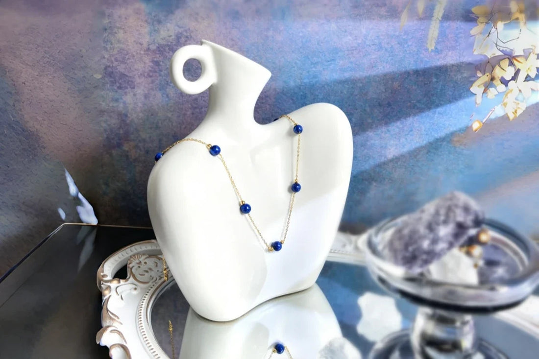 Blue lapis lazuli bead necklace on a delicate gold-tone chain, displayed on a white sculptural vase.
