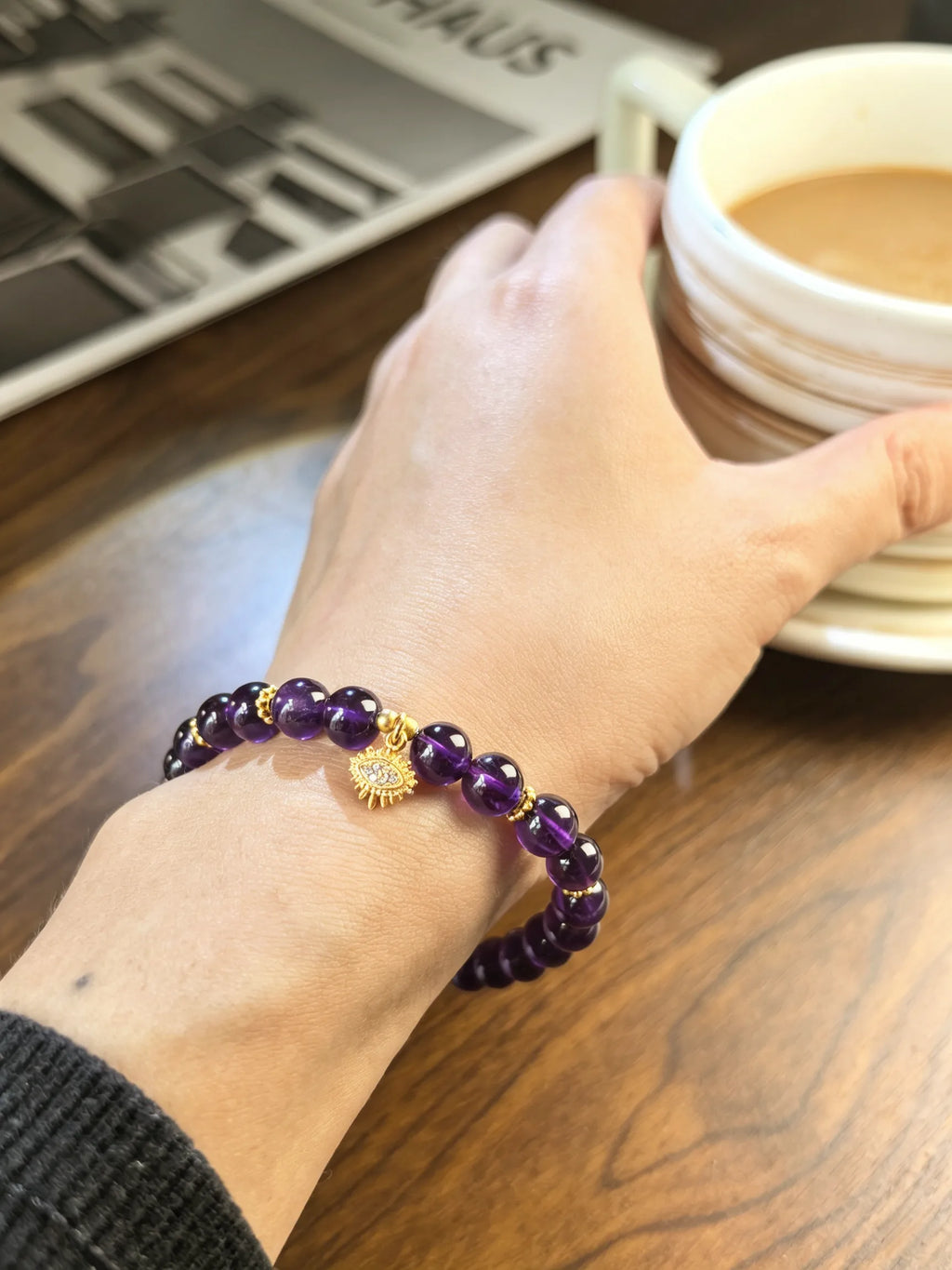 Purple amethyst bead bracelet with a gold-tone evil eye charm worn on a wrist beside a coffee cup.