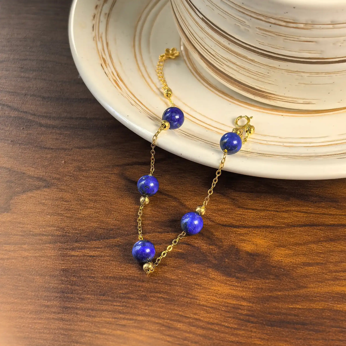 Blue lapis lazuli beads on a delicate gold-tone chain bracelet beside a ceramic coffee cup on a wooden table.