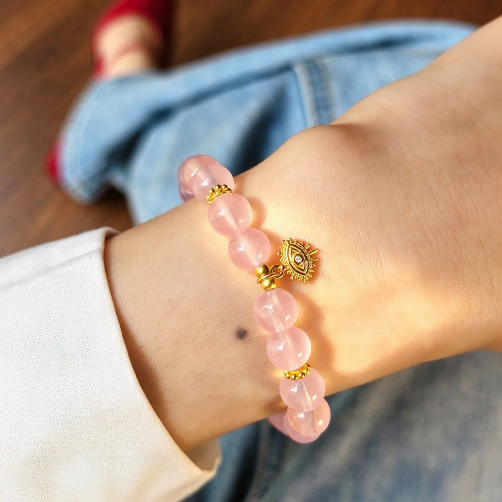 
Close-up of a rose quartz bracelet with gold-tone accents and an evil eye charm worn on a wrist.