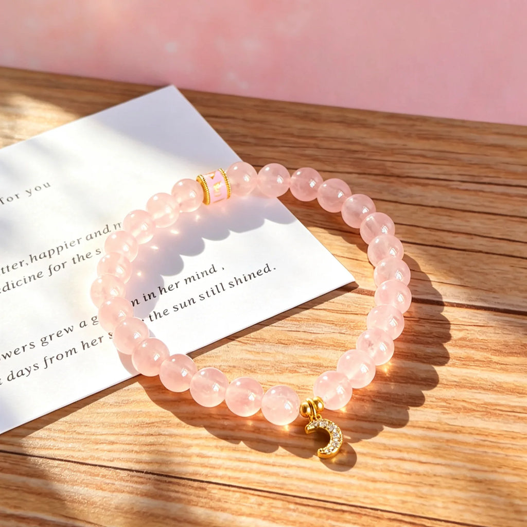 Pink rose quartz bracelet with a gold-tone evil eye charm on a wooden table beside a printed card.